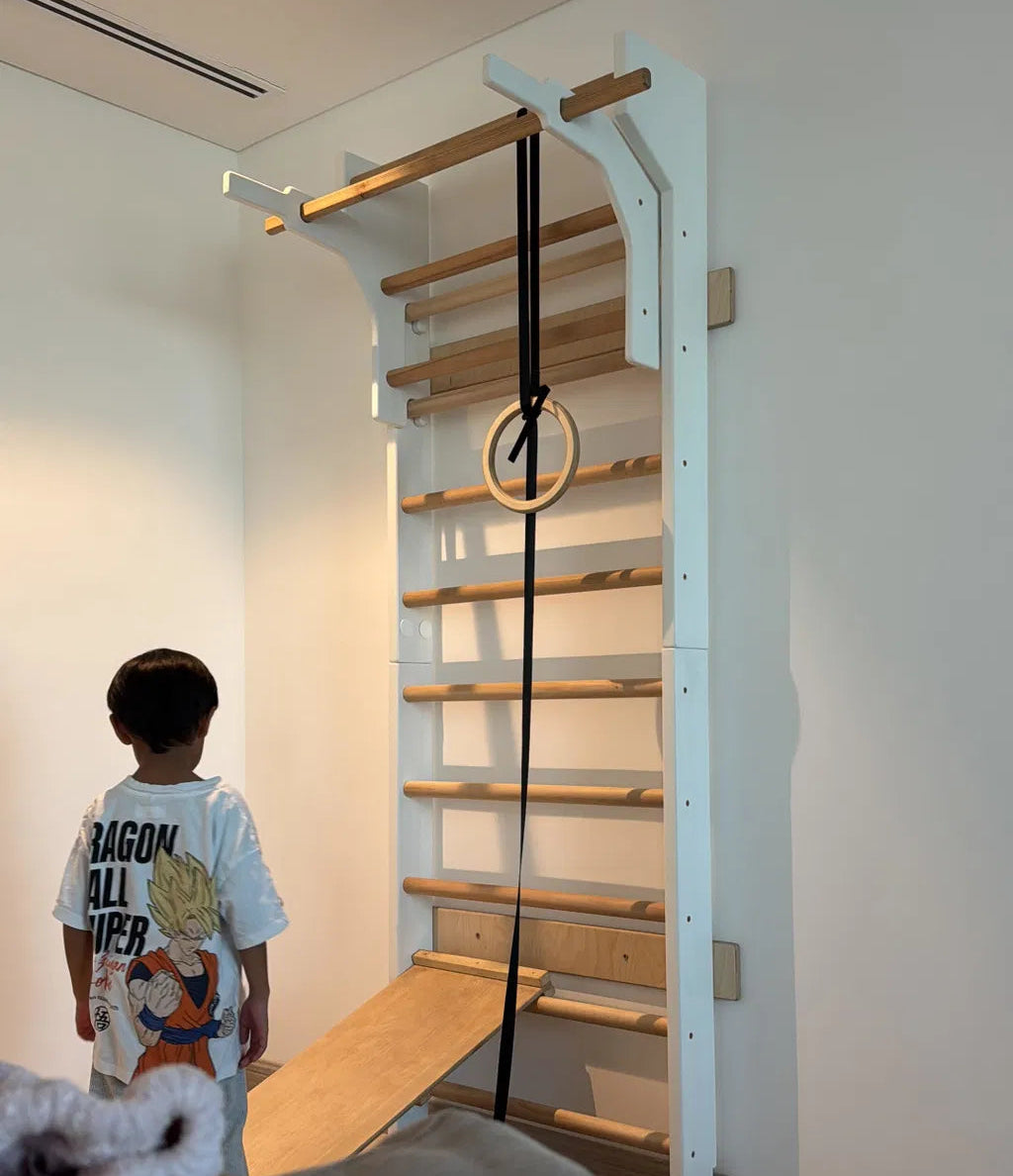 A child stands in a room looking at a wall-mounted wooden climbing ladder with white support brackets, a hanging black strap, and a wooden ring. A gray sofa is partially visible in the foreground.